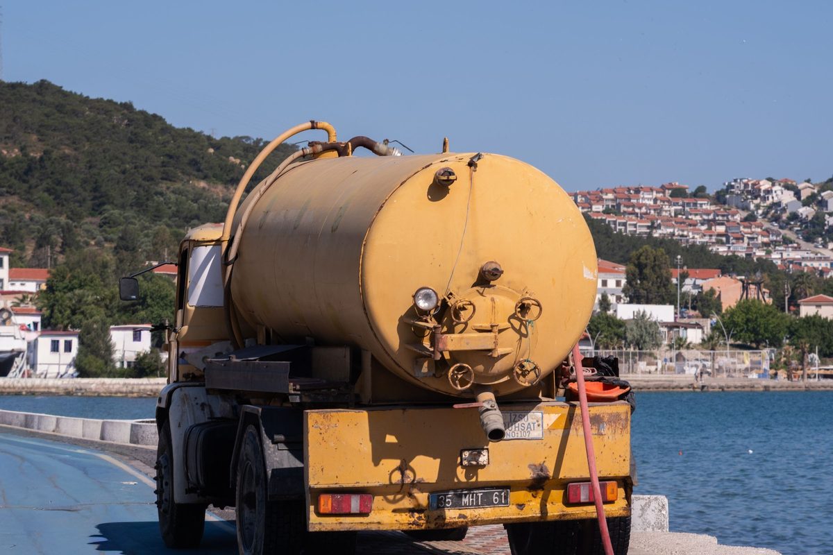 Yellow tank truck with a hose at the rear, parked by a harbor